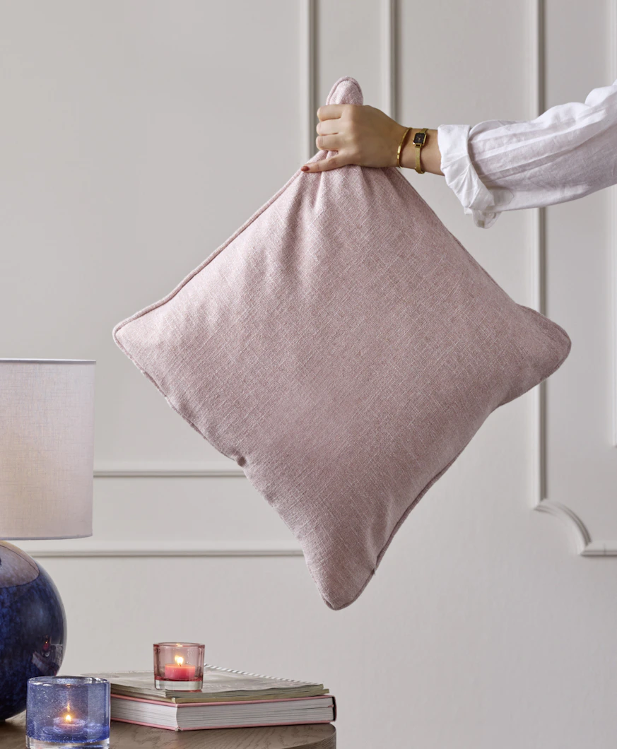 En hand håller i en prydnadskudde med rosa kuddfodral mot en vit vägg.