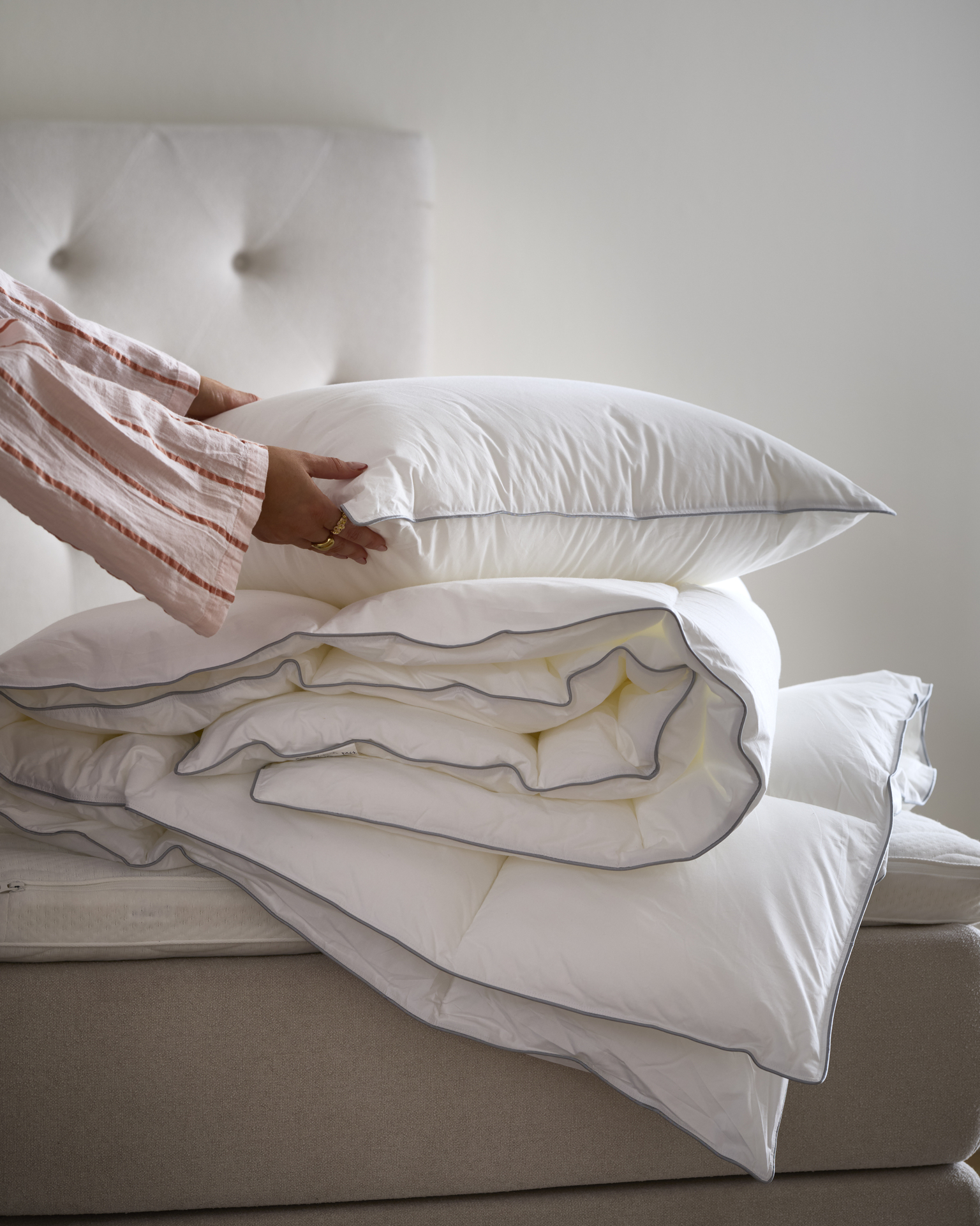 En hand håller i en kudde som ligger på ett hopvikt täcke på en obäddad säng.