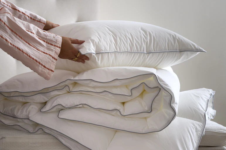 En hand håller i en kudde som ligger på ett hopvikt täcke på en obäddad säng.