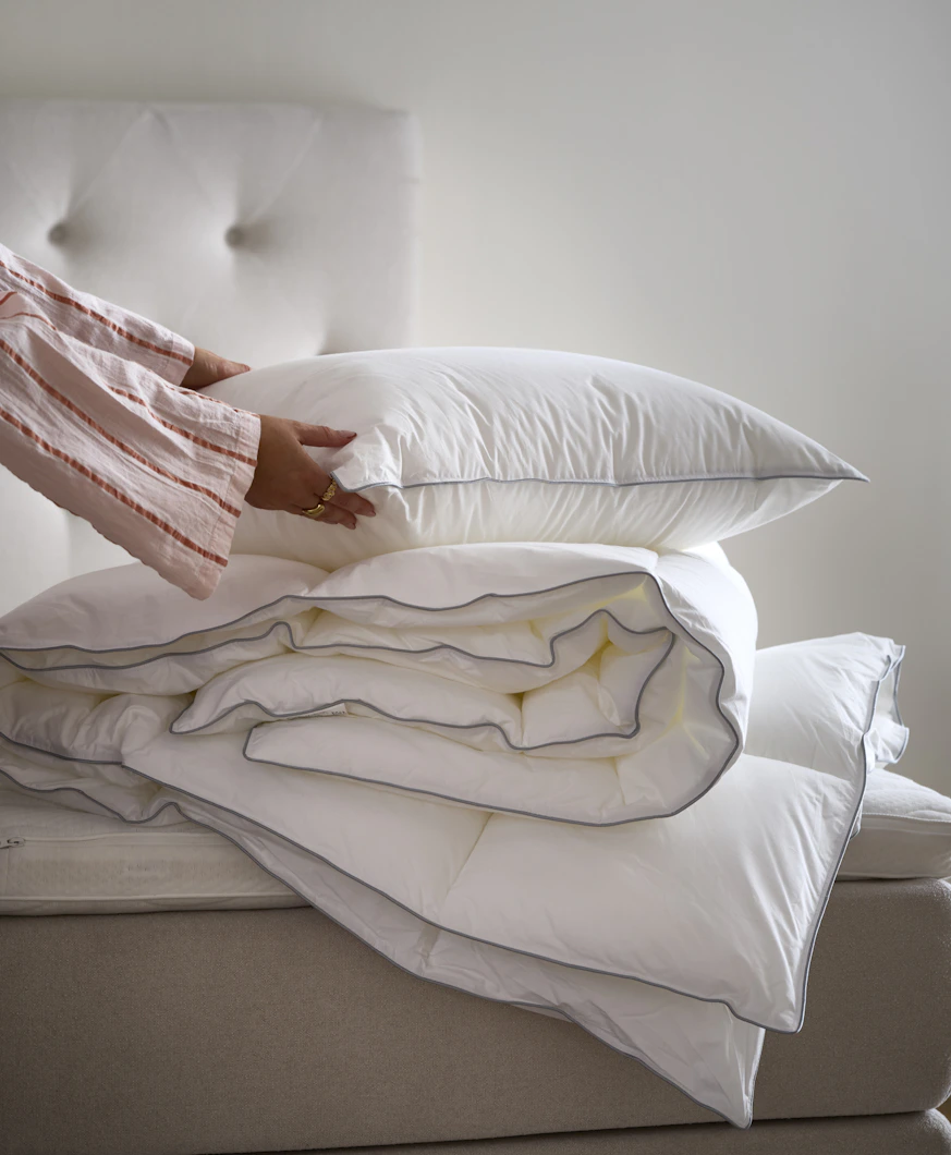 En hand håller i en kudde som ligger på ett hopvikt täcke på en obäddad säng.