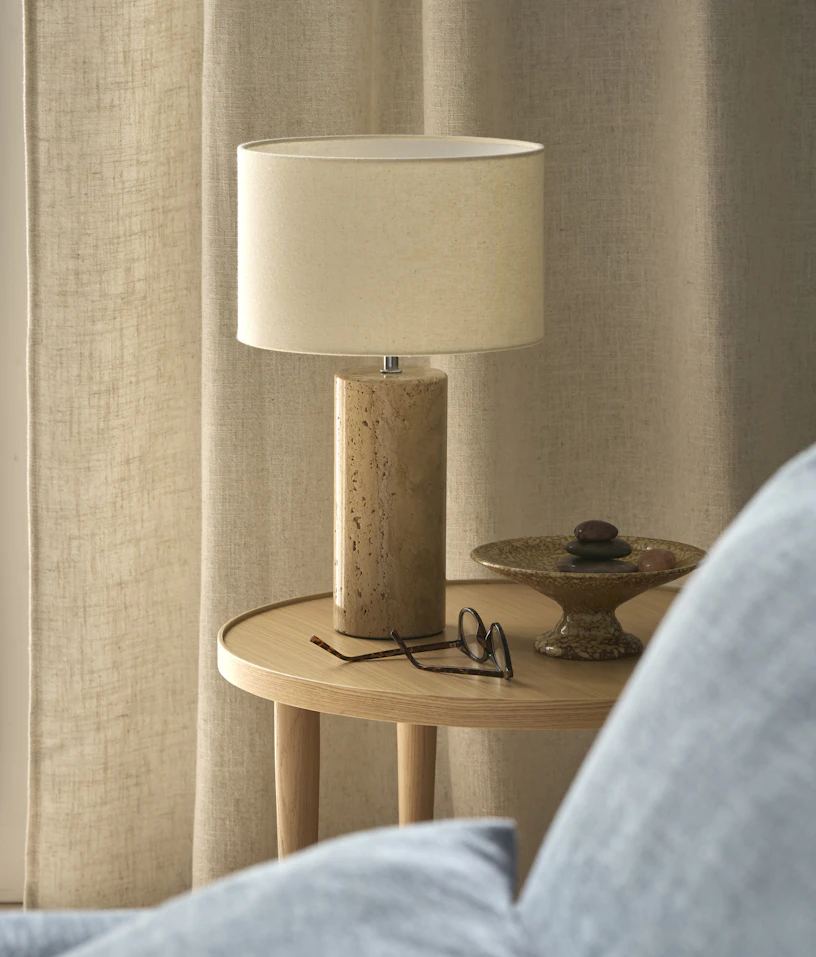 På ett sidobord i ek står en bordslampa i sandsten med vit lampskärm