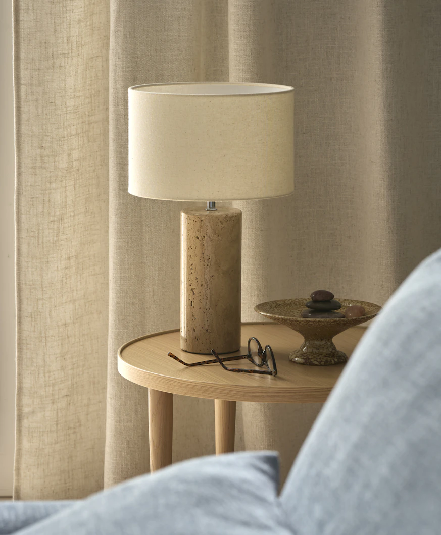 På ett sidobord i ek står en bordslampa i sandsten med vit lampskärm