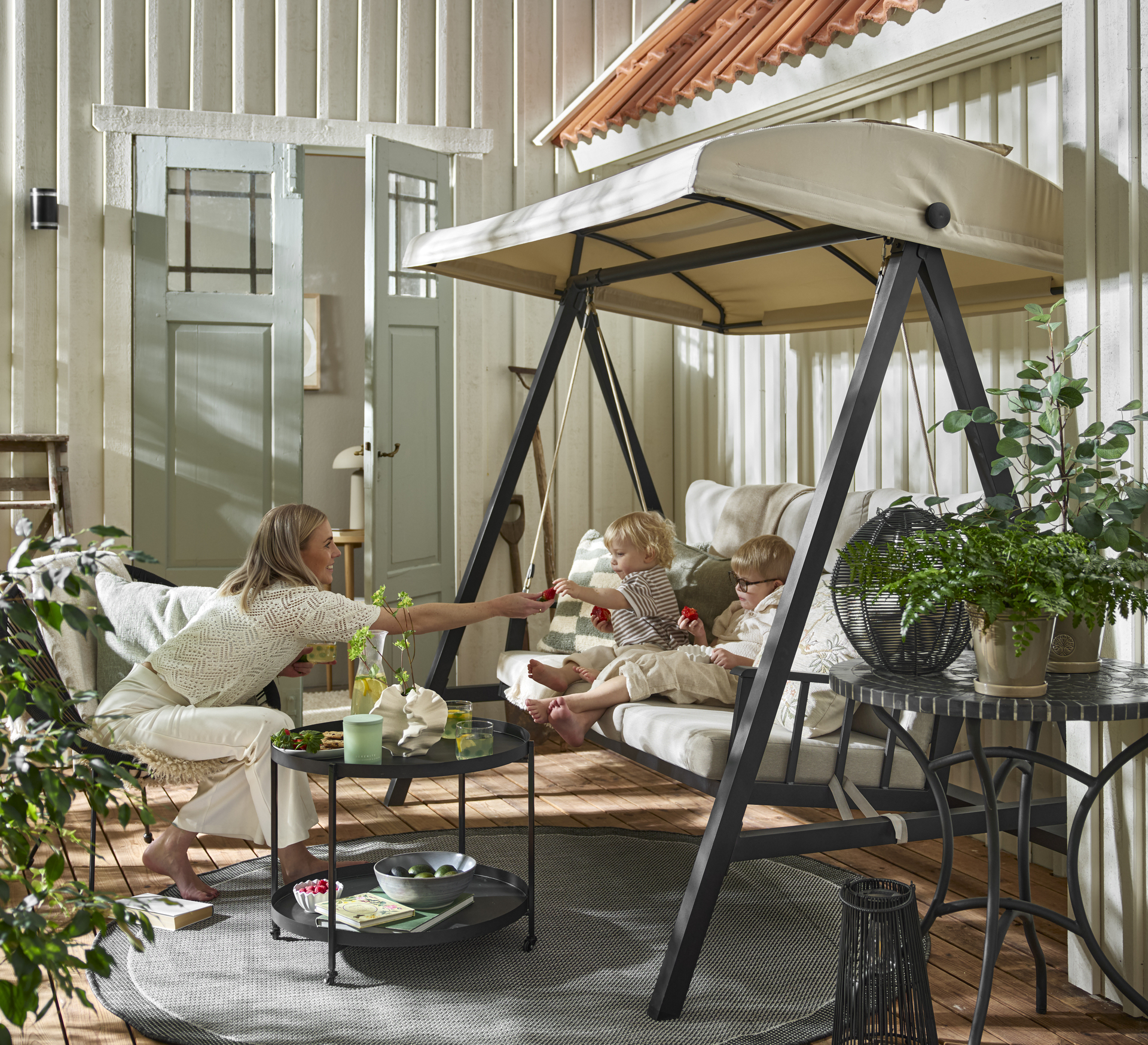 Två barn äter jordgubbar i en beige hammock med svart stomme. Mittemot sitter en kvinna i en utefåtölj. Uteplatsen är dekorerad med många växter.
