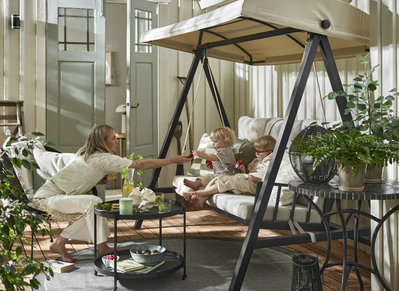 Två barn äter jordgubbar i en beige hammock med svart stomme. Mittemot sitter en kvinna i en utefåtölj. Uteplatsen är dekorerad med många växter.