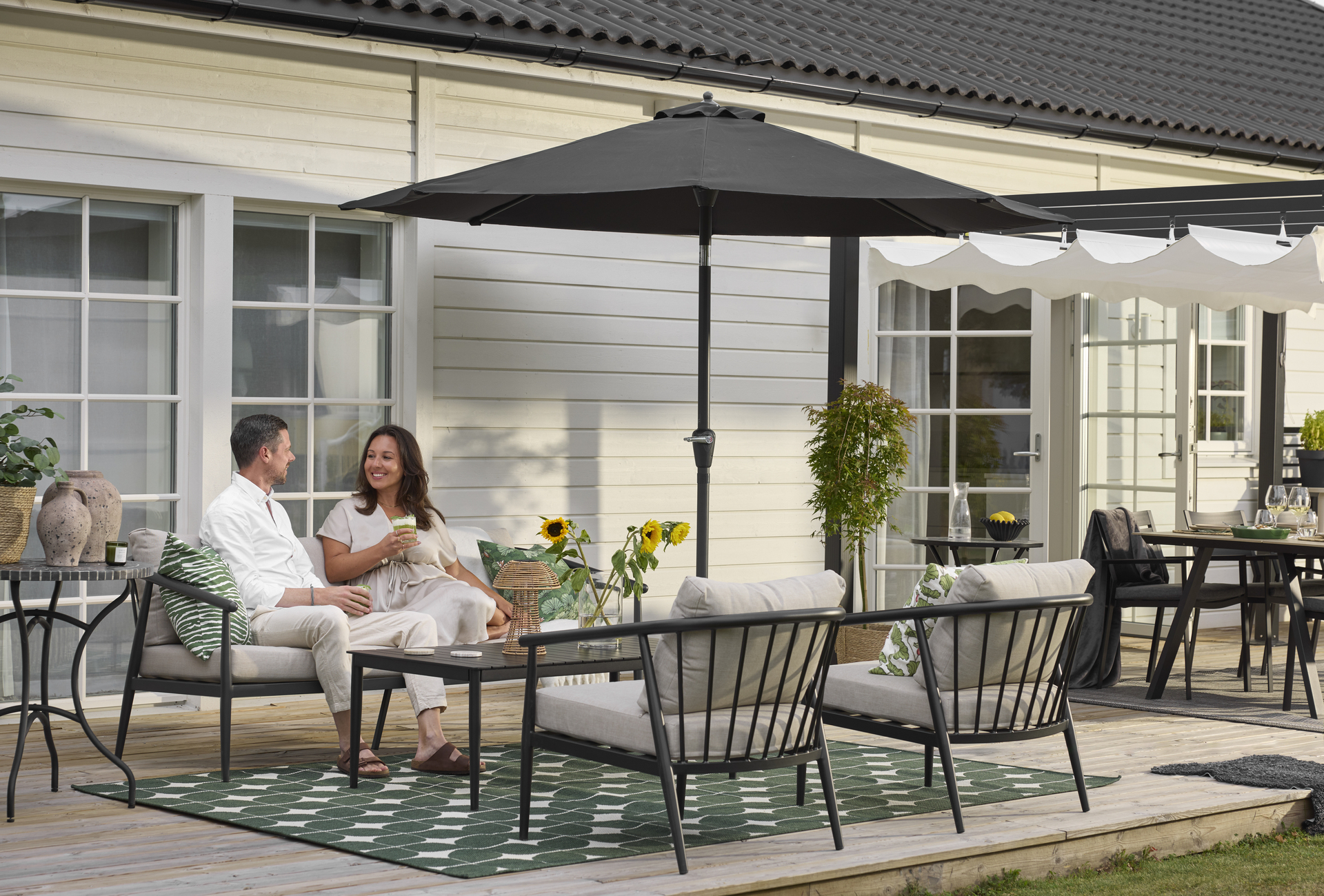 En man och en kvinna sitter i en svart utesoffa med beiga kuddar. Framför dem finns ett matchande soffbord och två fåtöljer. Under soffgruppen ligger en grönmönstrad utematta.