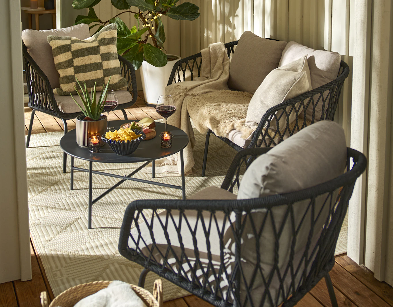 En mindre utesoffa med svart repflätning, ett svart soffbord och två matchande fåtöljer står på en uteplats. I soffan ligger plädar och i en av fåtöljerna ligger en grön och vitrutig kudde.