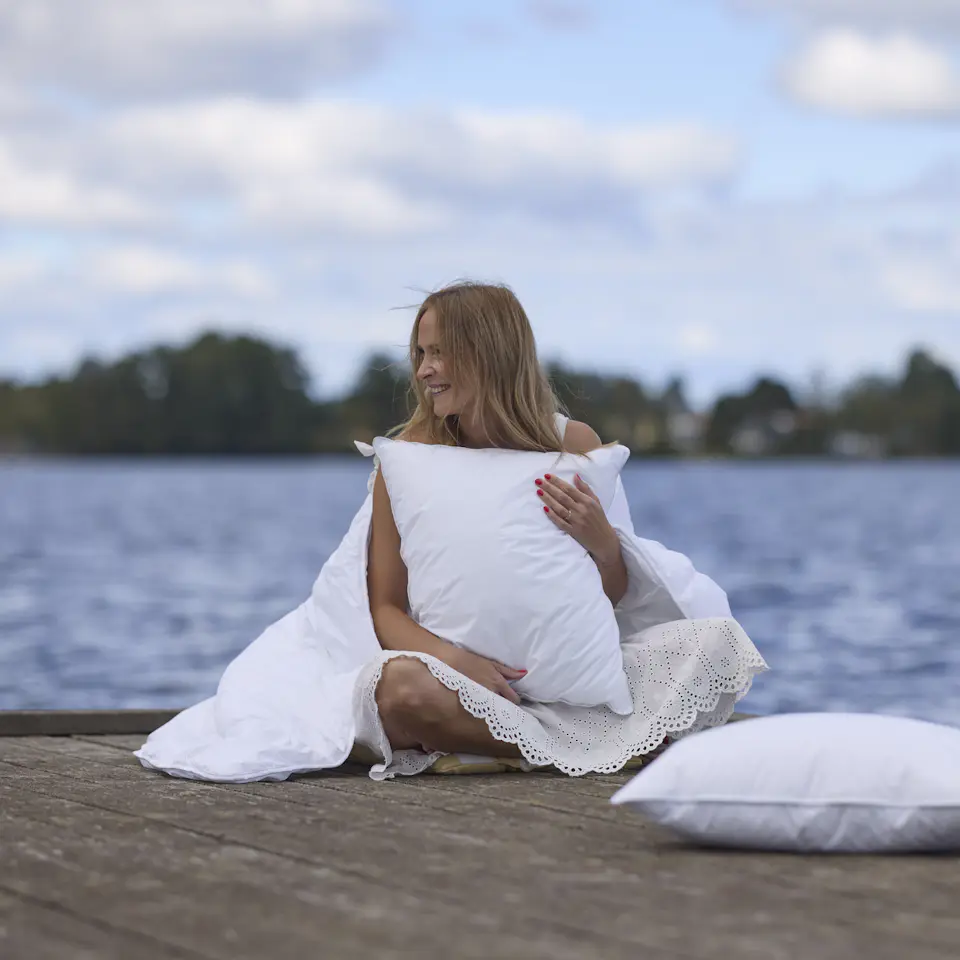 Blond kvinna sitter på brygga med vatten bakom och håller i vita kuddar