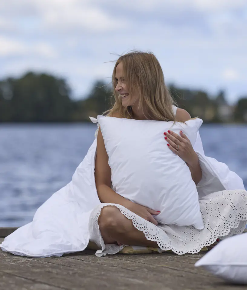 På en brygga sitter en kvinna invirad i ett täcke. I famnen håller hon en kudde.