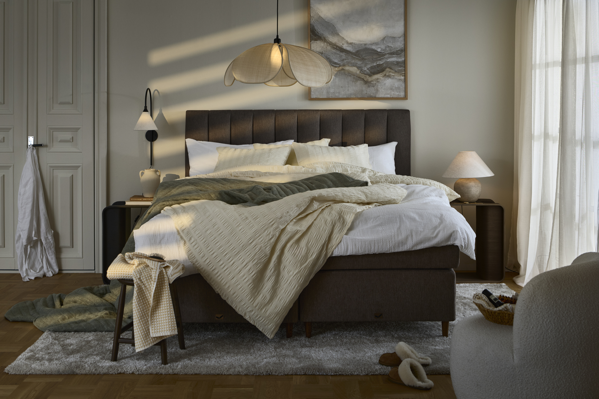 Sovrum i skymningsljus med stor kontinentalsäng i brunt tyg och sänggavel centralt placerad i rummet, sängen är bäddad med sängkläder i nyansera av beige, vitt och grönt.