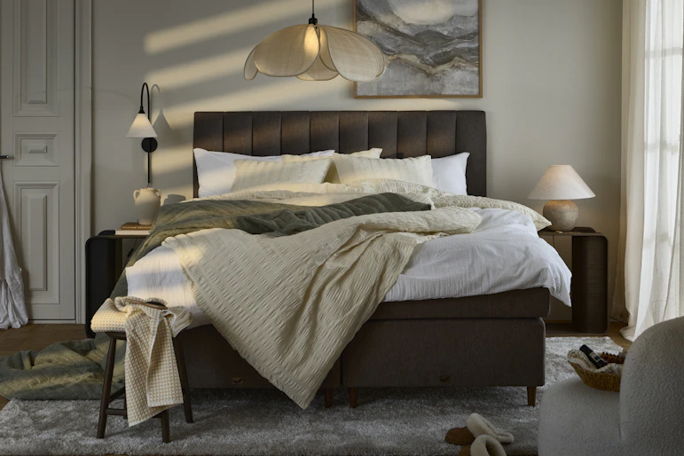 Sovrum i skymningsljus med stor kontinentalsäng i brunt tyg och sänggavel centralt placerad i rummet, sängen är bäddad med sängkläder i nyansera av beige, vitt och grönt.