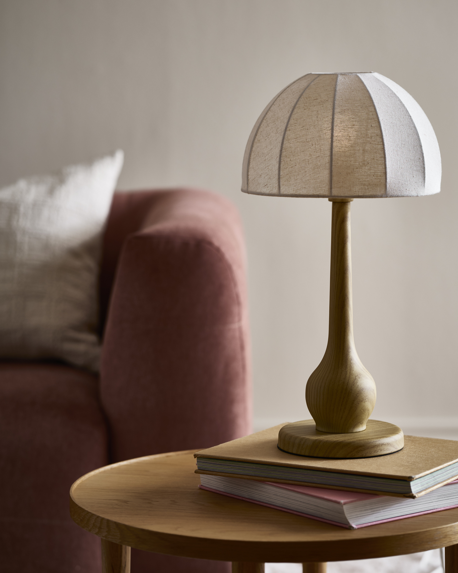 En bordslampa med vit linneskärm och träfärgad fot står på ett bord framför en rosa soffa.