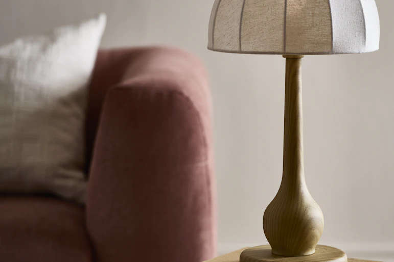 En bordslampa med vit linneskärm och träfärgad fot står på ett bord framför en rosa soffa.