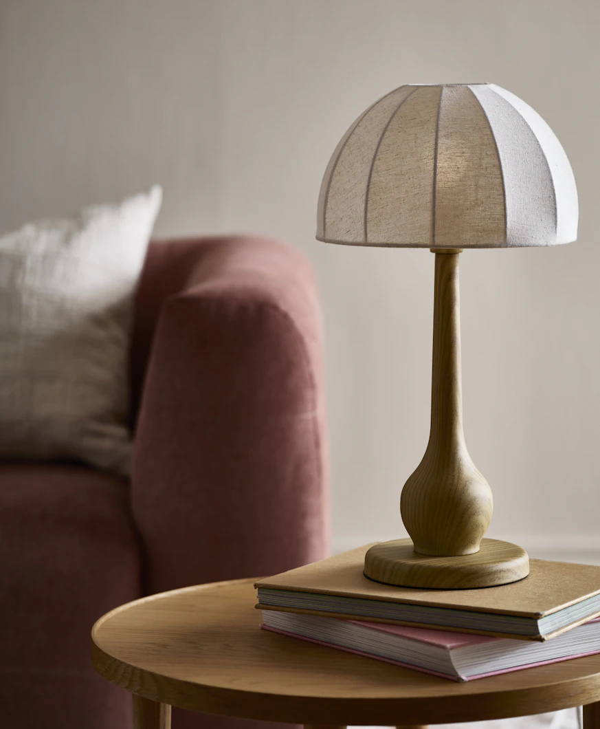 En bordslampa med vit linneskärm och träfärgad fot står på ett bord framför en rosa soffa.