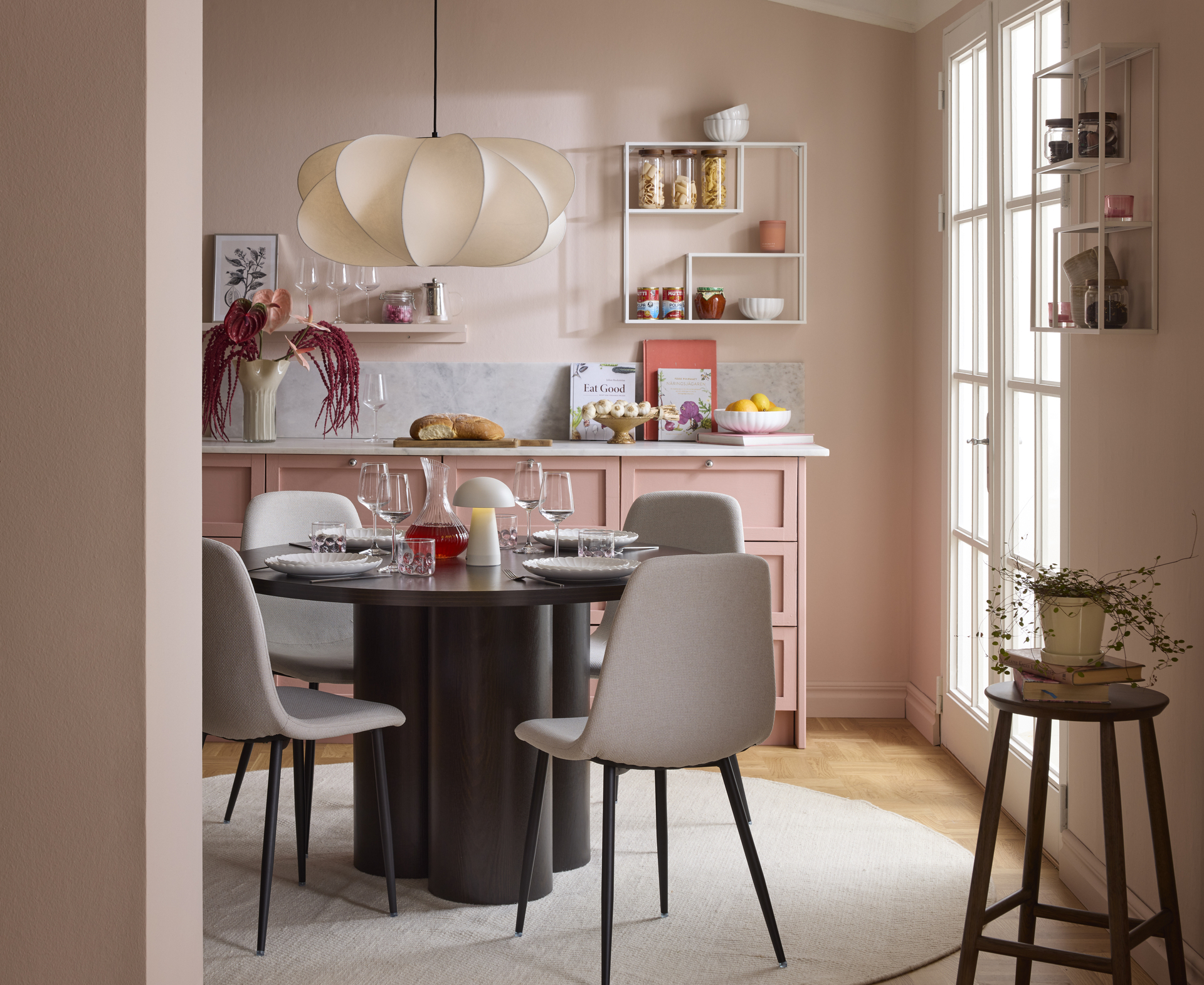 Matbord med kraftig bas av tre cylindrar och rund bordsskiva i mörkt trä samt fyra stolar runtom med ljusbeige klädsel och mörka ben står i ett kök med rosa skåp och väggar.