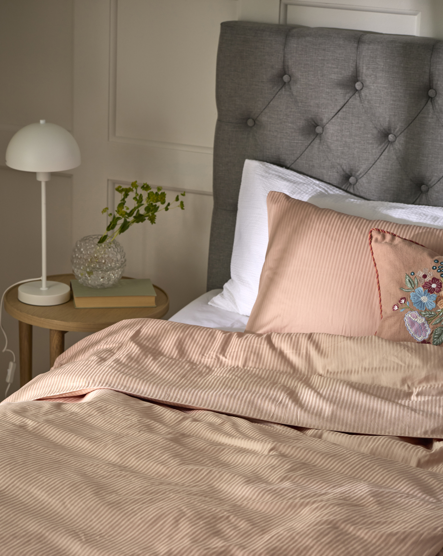 En säng med grå pikerad sänggavel är bäddad med randiga sängkläder i rosa.