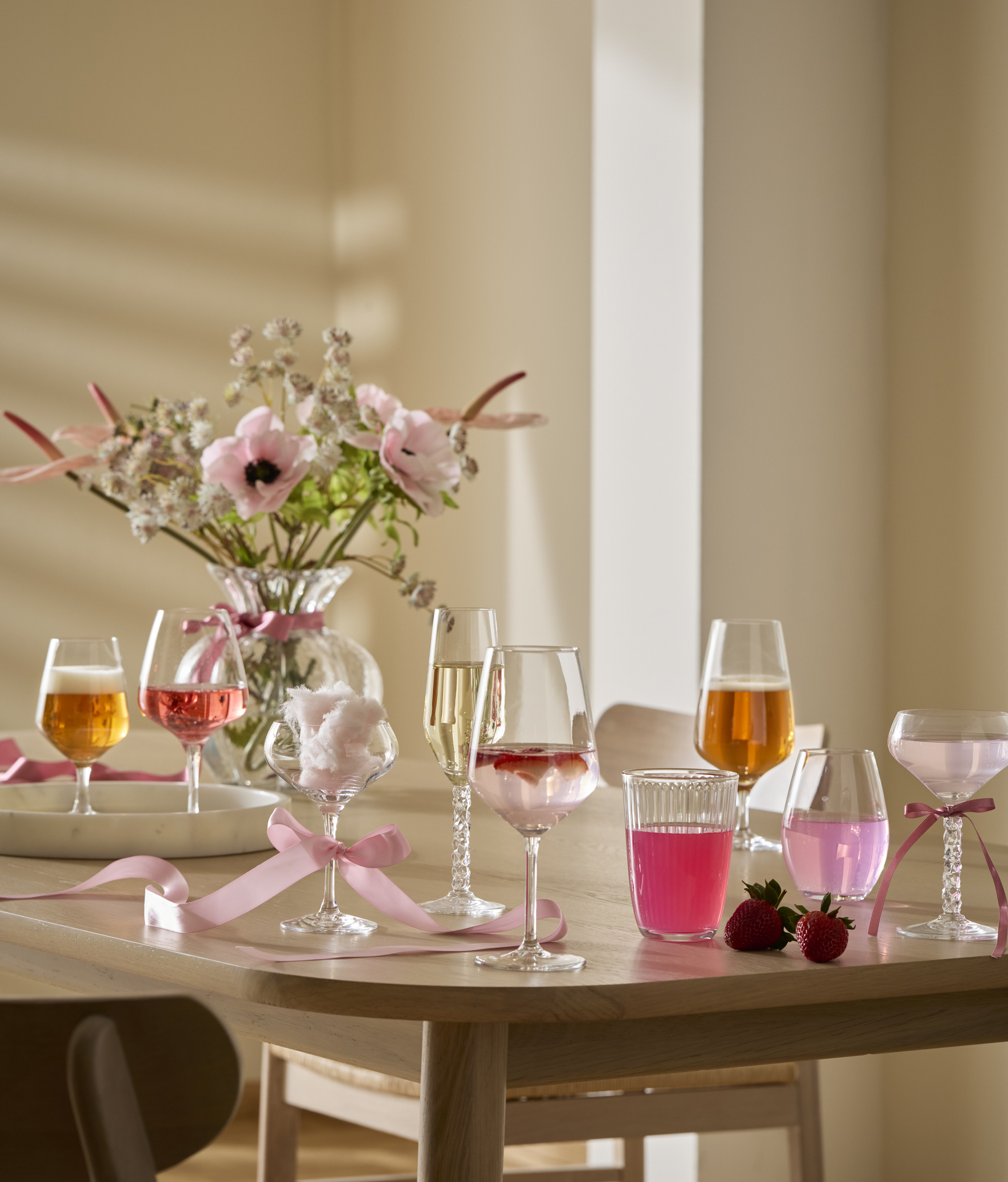 Festliga glas i olika modeller och en vas med rosa blommor står på ett ljust träbord. 