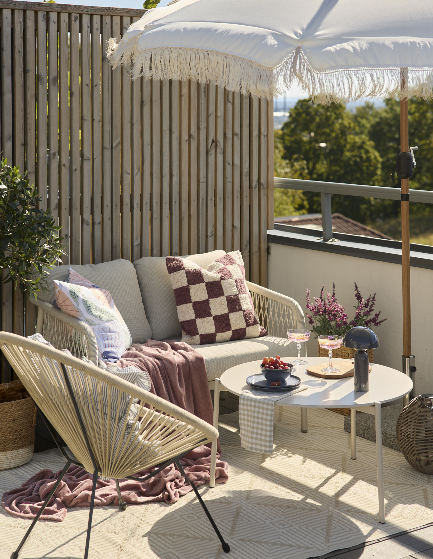 En 2-sitssoffa och en utefåtölj står under ett parasoll på en balkong. I soffan ligger en rutig kudde i vitt och mörkrosa och en matchande mörkrosa pläd.
