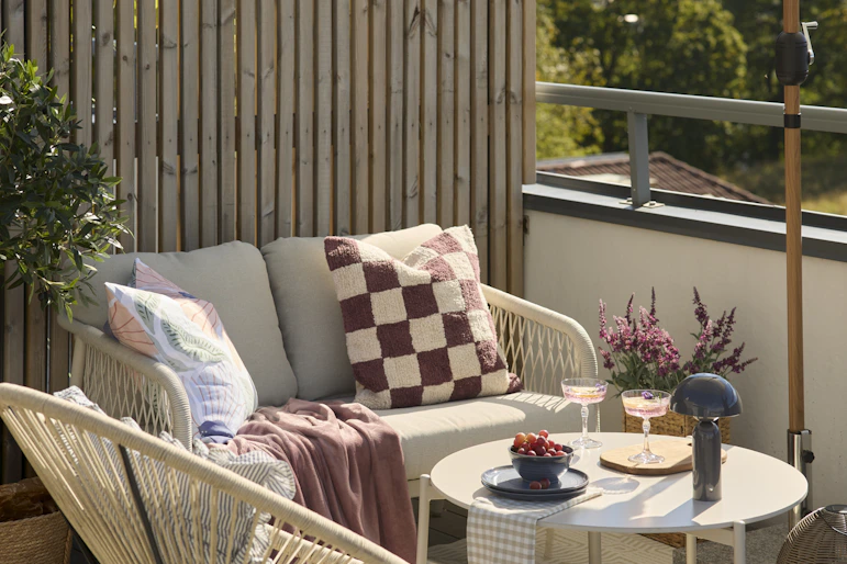 En 2-sitssoffa och en utefåtölj står under ett parasoll på en balkong. I soffan ligger en rutig kudde i vitt och mörkrosa och en matchande mörkrosa pläd.