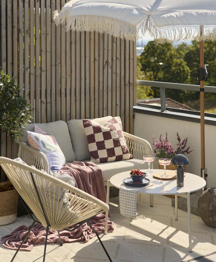 En 2-sitssoffa och en utefåtölj står under ett parasoll på en balkong. I soffan ligger en rutig kudde i vitt och mörkrosa och en matchande mörkrosa pläd.
