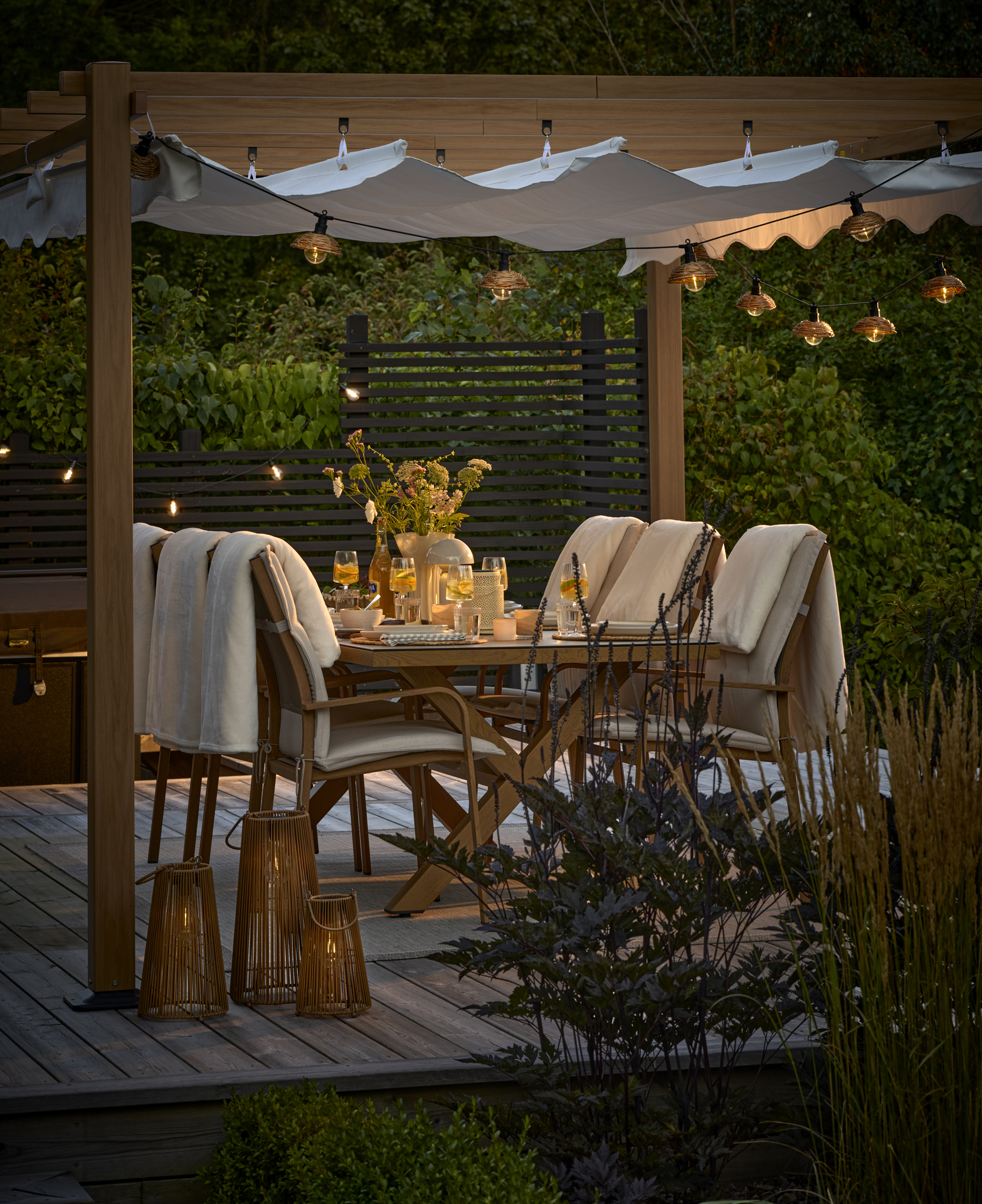 En uteplats med matgrupp och pergola i skymning. Uteplatsen är belyst med ljusslinga och solcellslyktor.