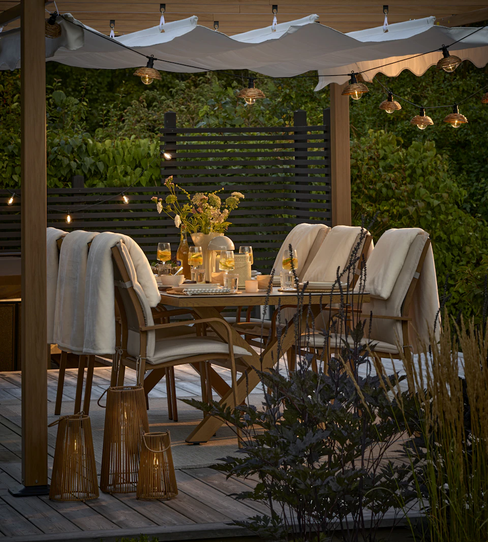 En uteplats med matgrupp och pergola i skymning. Uteplatsen är belyst med ljusslinga och solcellslyktor.