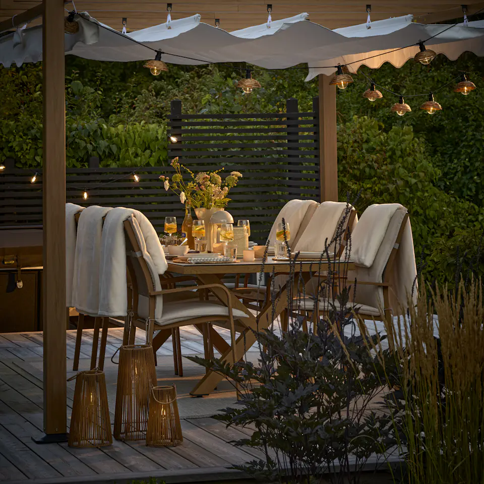 En uteplats med matgrupp och pergola i skymning. Uteplatsen är belyst med ljusslinga och solcellslyktor.