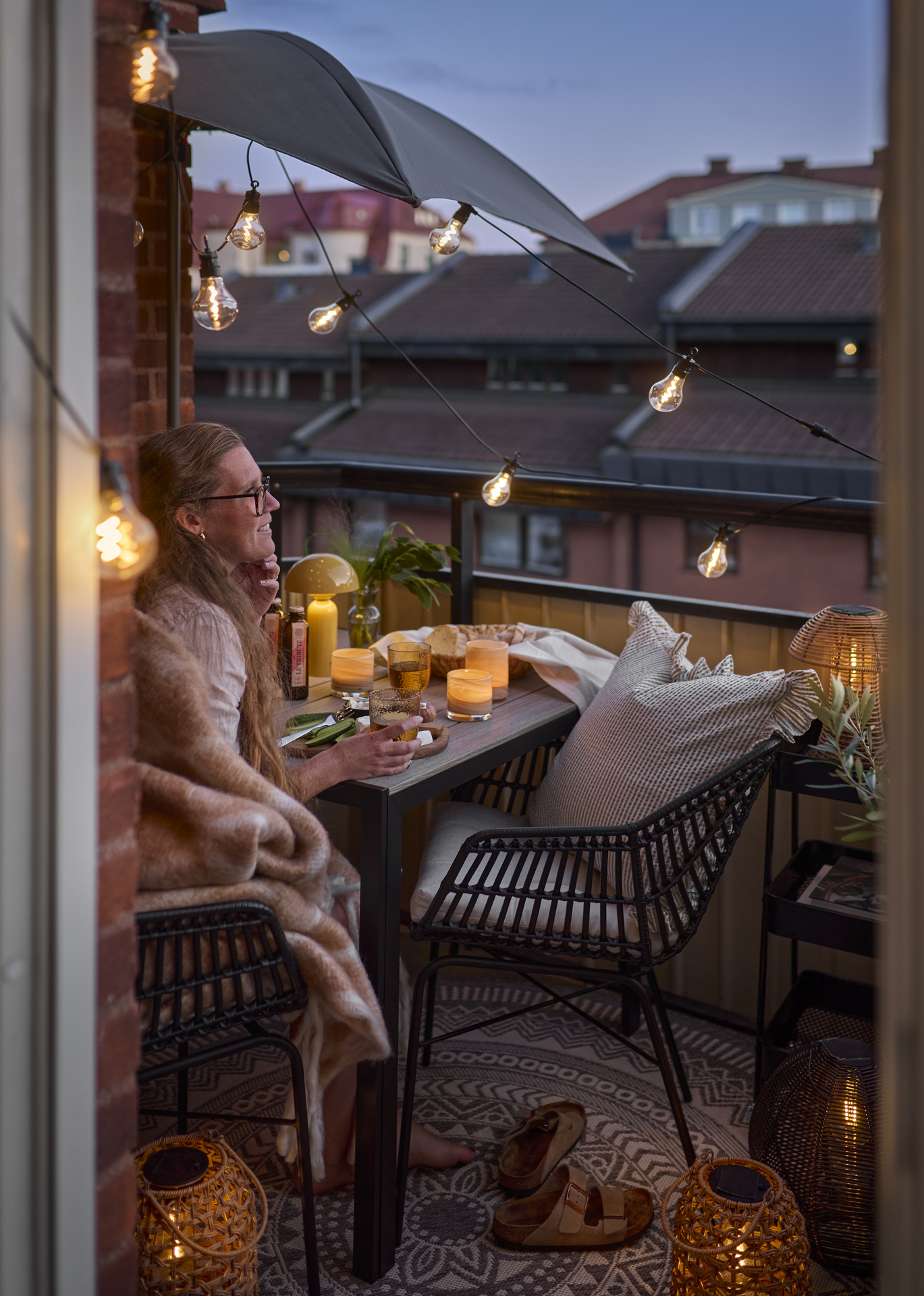 En tjej sitter på sin balkong i skymning och blickar ut över takåsarna. Balkongen är belyst med ljusslinga och portabel bordslampa.