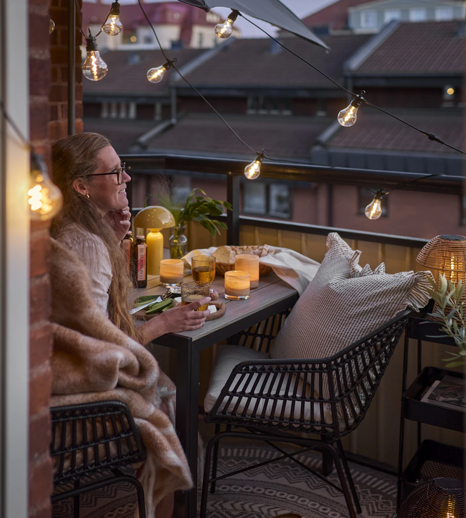 En tjej sitter på sin balkong i skymning och blickar ut över takåsarna. Balkongen är belyst med ljusslinga och portabel bordslampa.