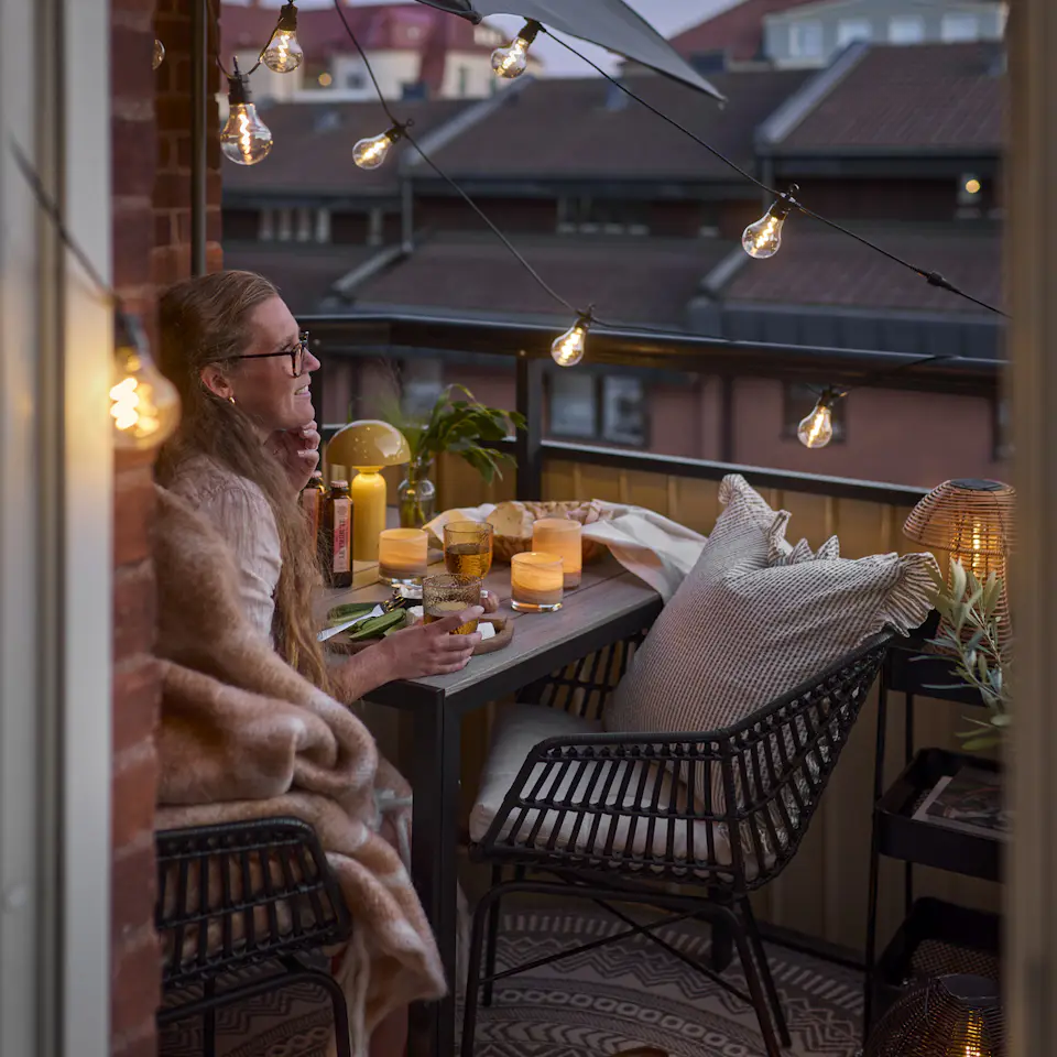 En tjej sitter på sin balkong i skymning och blickar ut över takåsarna. Balkongen är belyst med ljusslinga och portabel bordslampa.