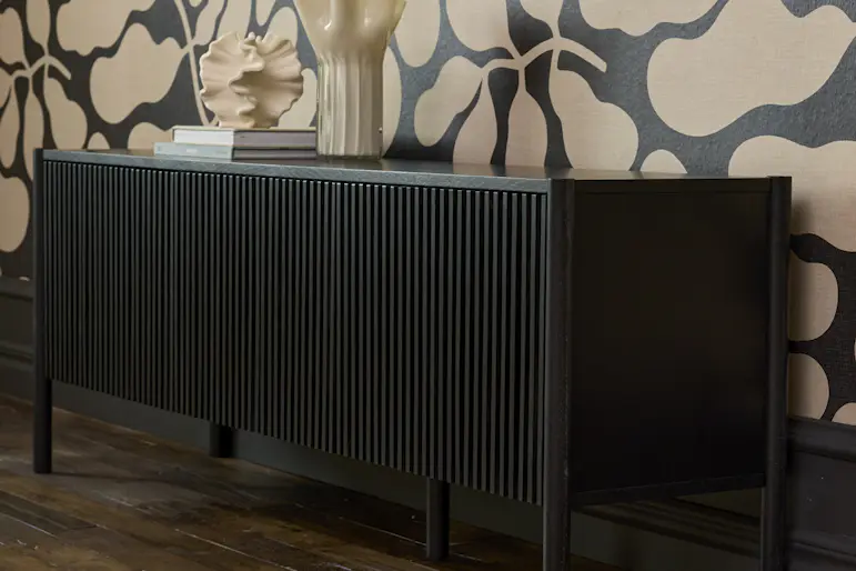 Svartlackerad tv-bänk med ribbad front står mot en vägg med blåmönstrad tapet.