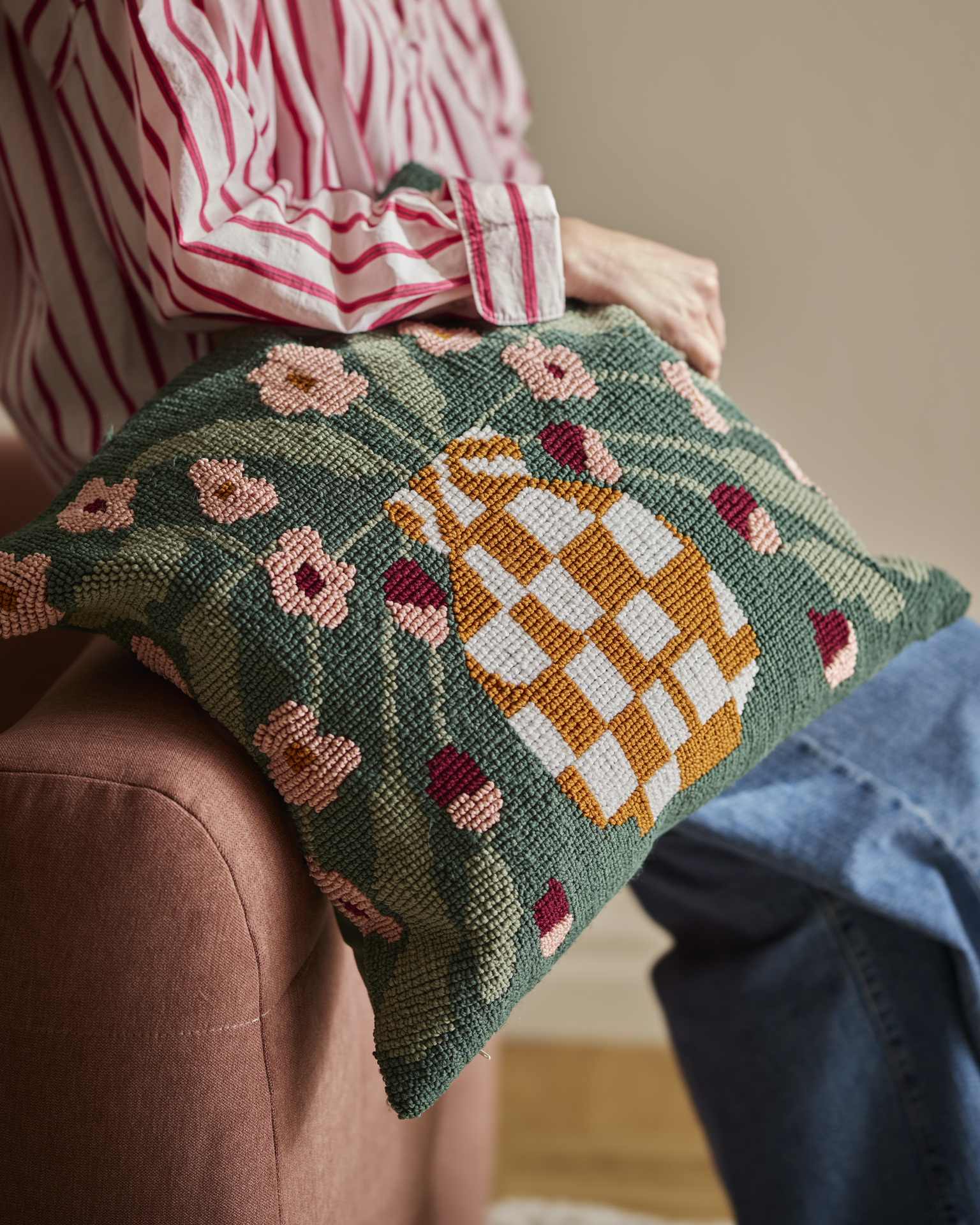 En person håller i en prydnadskudde med stickad struktur och dekorativt mönster av en vas fylld med blommor. 