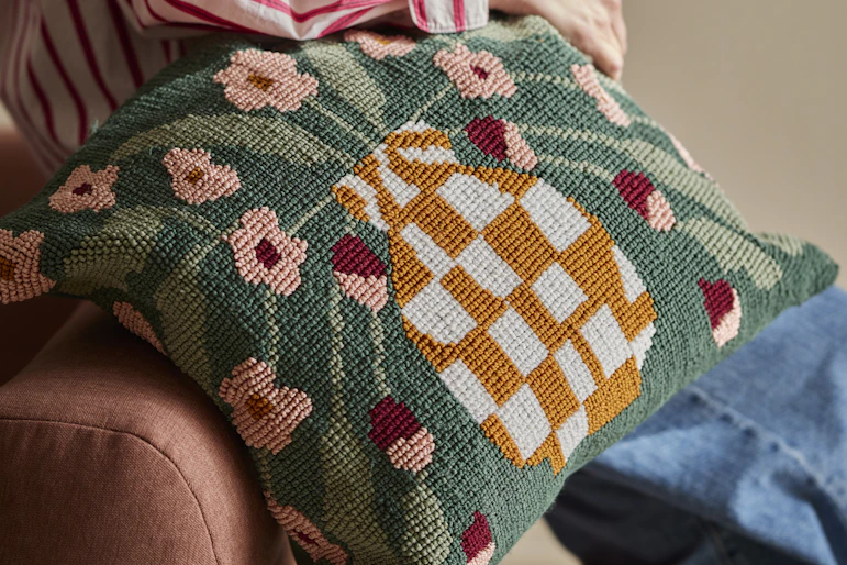 En person håller i en prydnadskudde med stickad struktur och dekorativt mönster av en vas fylld med blommor.