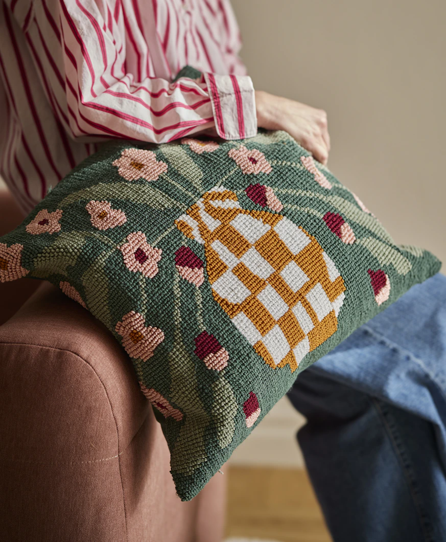 En person håller i en prydnadskudde med stickad struktur och dekorativt mönster av en vas fylld med blommor.