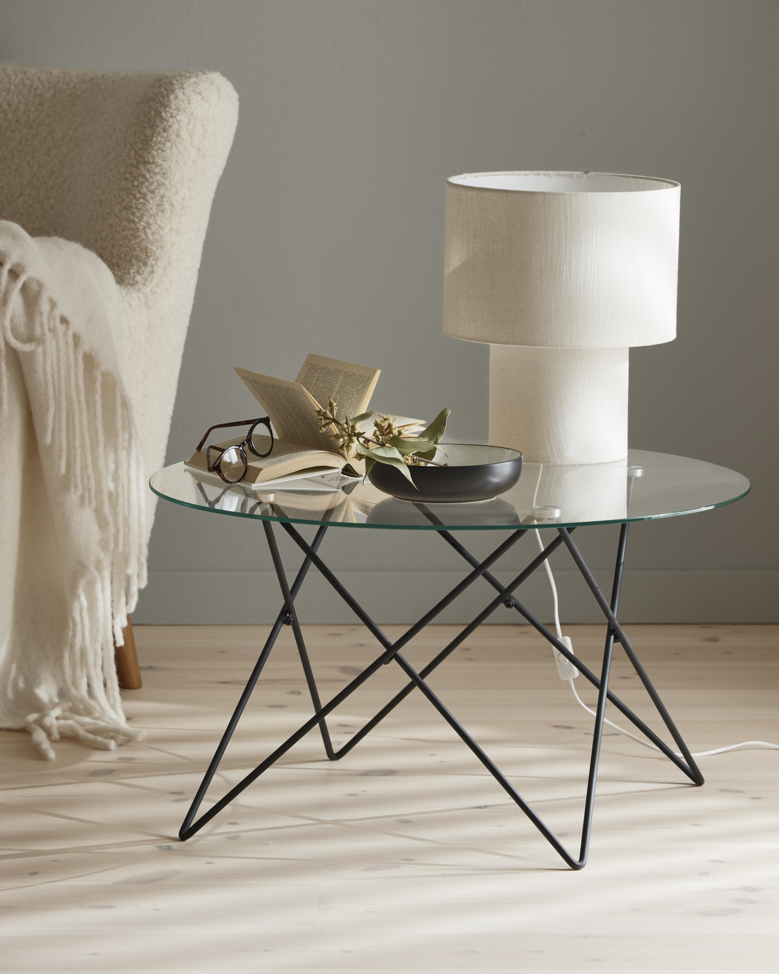 En vit lampa, en bok och en skål står på ett runt soffbord med glasskiva och underrede av metall.