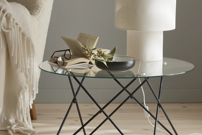 En vit lampa, en bok och en skål står på ett runt soffbord med glasskiva och underrede av metall.