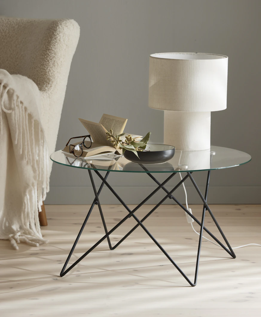 En vit lampa, en bok och en skål står på ett runt soffbord med glasskiva och underrede av metall.