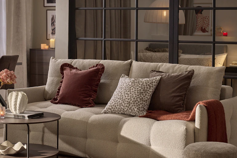 En beige bäddsoffa med rostfärgade och mönstrade kuddar står i ett dovt belyst rum.