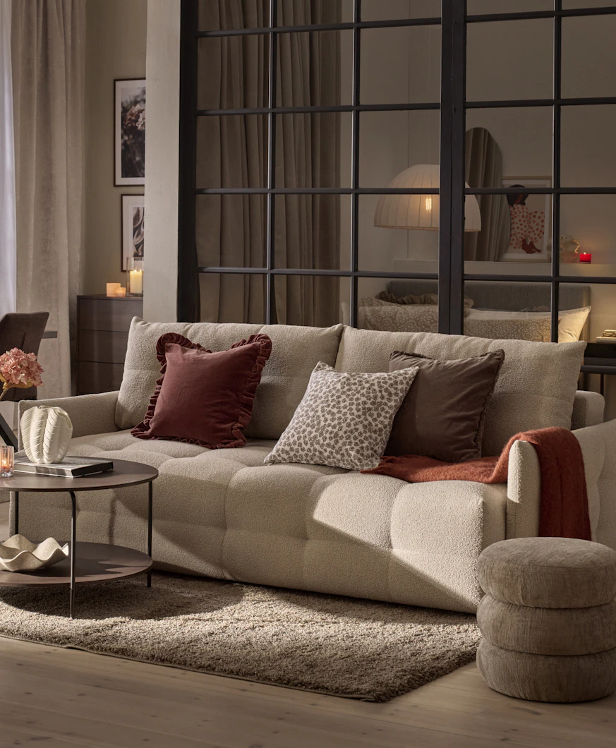 En beige bäddsoffa med rostfärgade och mönstrade kuddar står i ett dovt belyst rum.