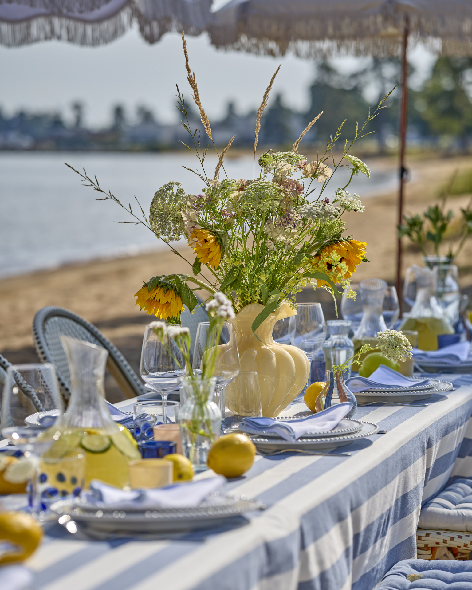 Detaljbild på långbord som står på stranden med gul vas, blå- och vitrandig duk och somrigt porslin.
