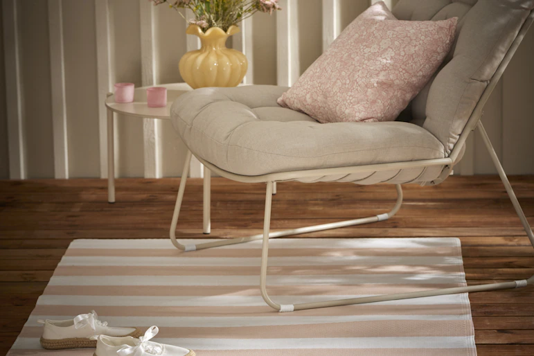 En ljus utefåtölj med rosa kudde står på en rosa- och vitrandig utematta-