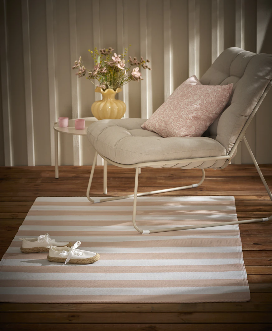 En ljus utefåtölj med rosa kudde står på en rosa- och vitrandig utematta-