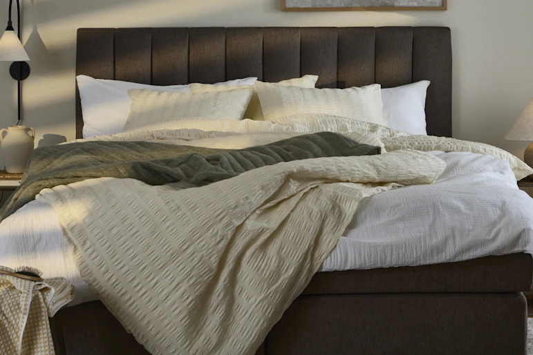 I ett dovt belyst sovrum står en mörkbrun säng bäddad med neutrala textilier