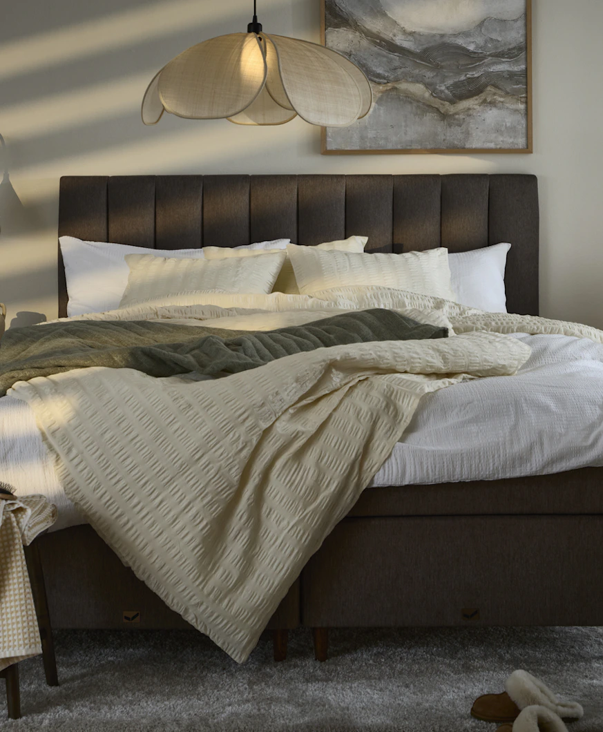 I ett dovt belyst sovrum står en mörkbrun säng bäddad med neutrala textilier