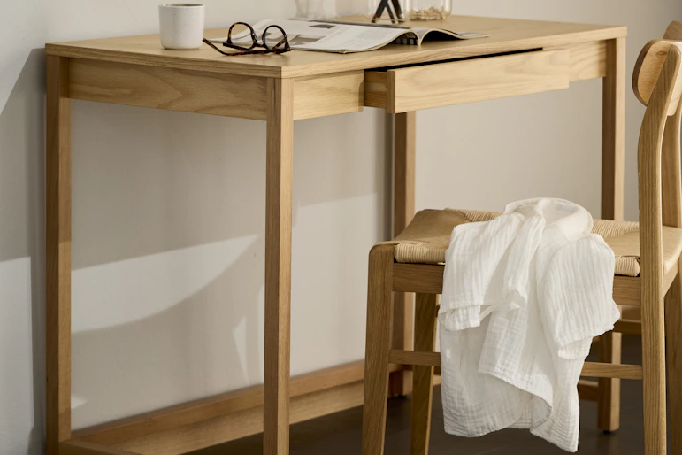 Ett skrivbord i ljust trä och matchande stol står framför en ljus vägg.