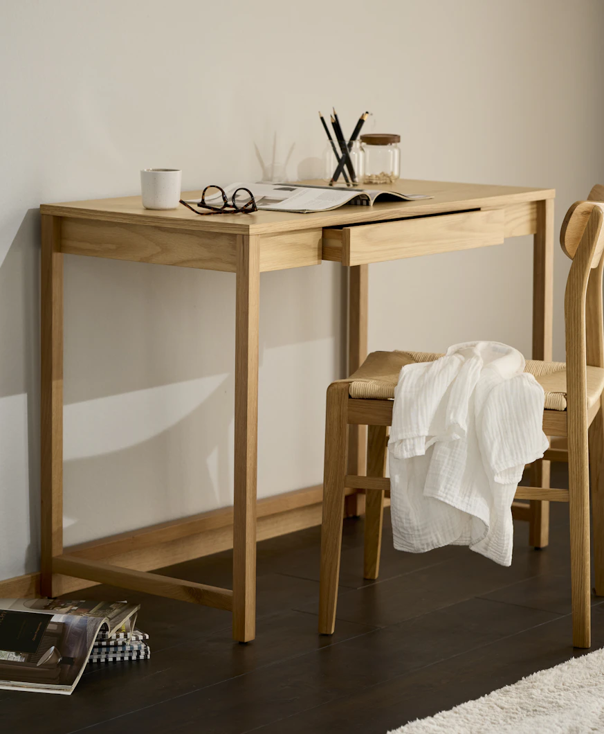 Ett skrivbord i ljust trä och matchande stol står framför en ljus vägg.