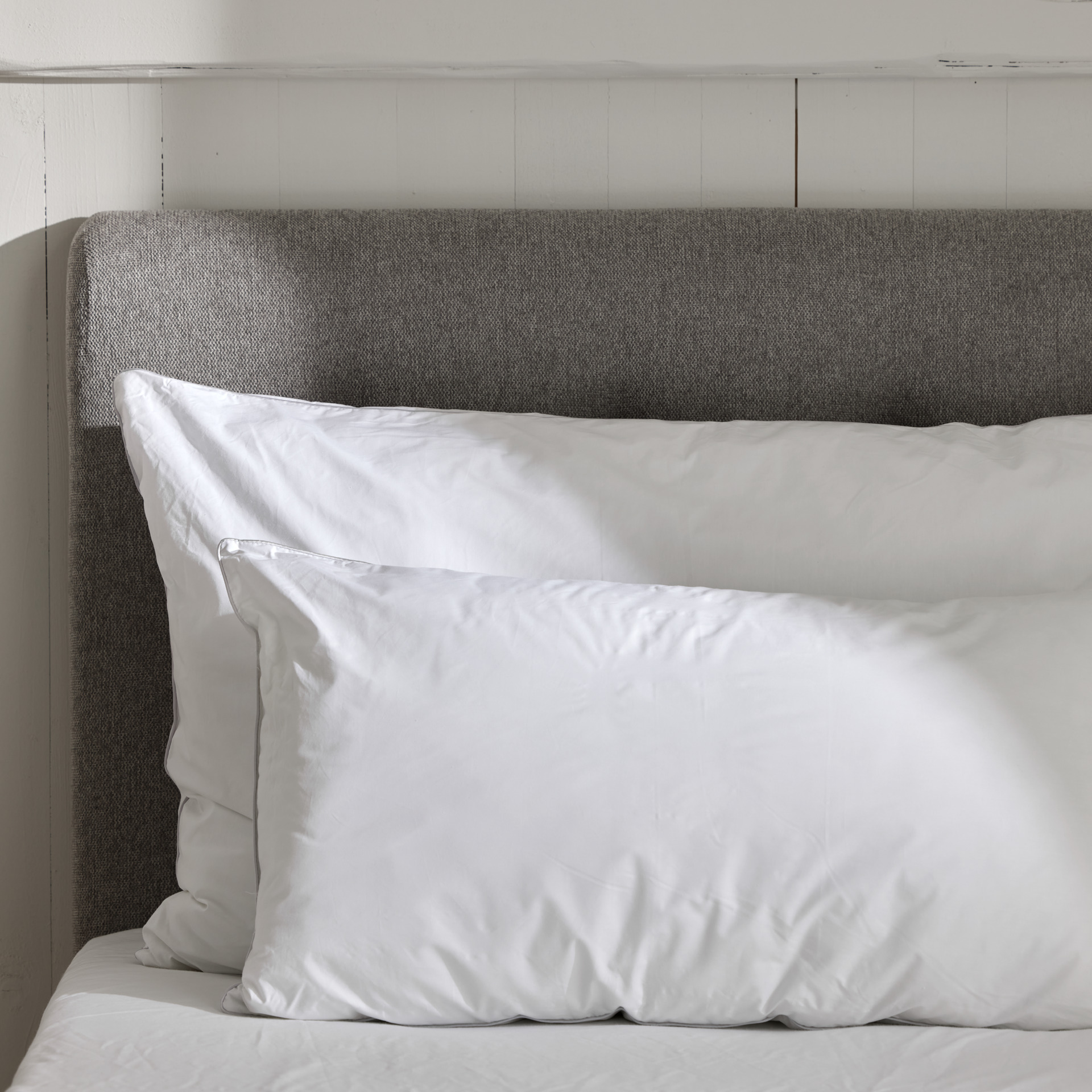 Två hotellkuddar i olika storlekar ligger framför en grå sänggavel.