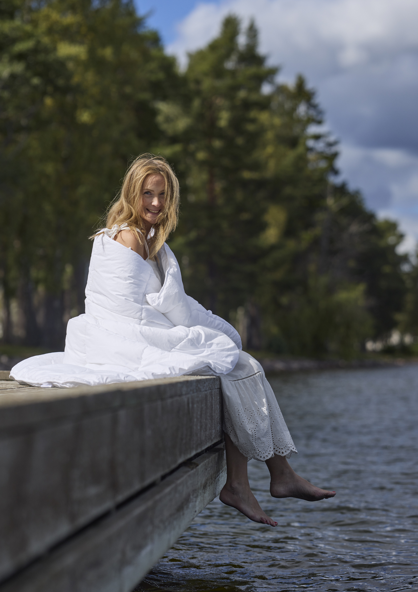 En kvinna invirad i ett täcke utan påslakan sitter på en brygga i sommaren.
