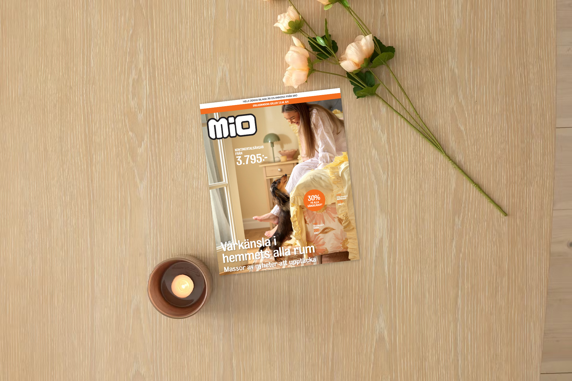 På ett ljust träbord ligger kampanjbladet för Mios vårmyskampanj intill ett ljus och en blomma.