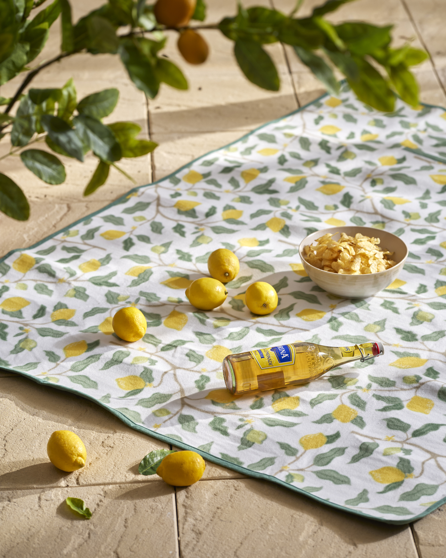 en picknickfilt med citronmotiv ligger på ett golv och på filten finns en skål med chips, en glasflaska och citroner