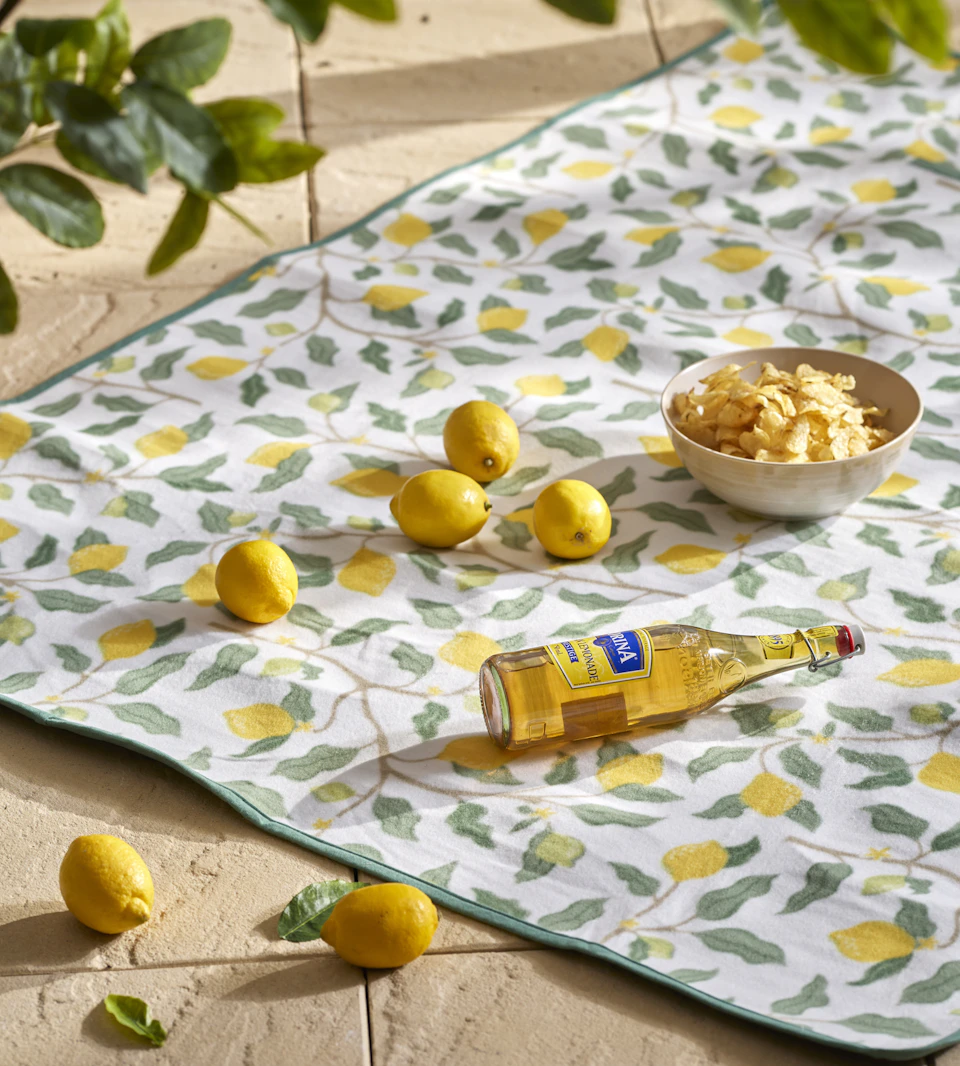 en picknickfilt med citronmotiv ligger på ett golv och på filten finns en skål med chips, en glasflaska och citroner